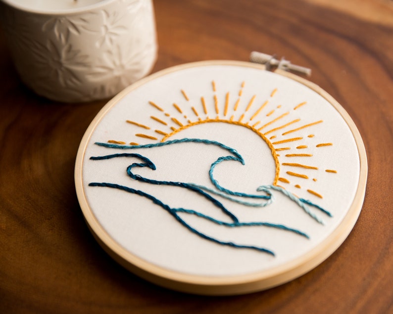 Peut inclure: Art de broderie en forme de cercle repr&eacute;sentant une vague bleue avec un soleil jaune qui se l&egrave;ve au-dessus.