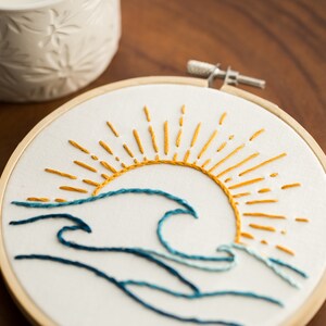 Peut inclure: Art de broderie en forme de cercle repr&eacute;sentant un soleil stylis&eacute; avec des rayons jaunes et une vague bleue. Le soleil se couche derri&egrave;re la vague, cr&eacute;ant une sc&egrave;ne calme et sereine.