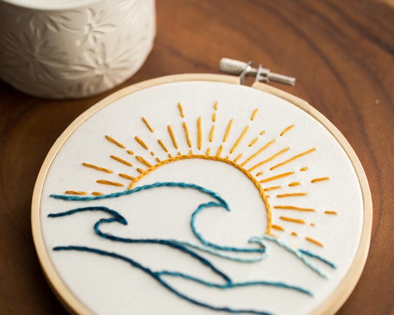 Peut inclure: Art de broderie en forme de cercle repr&eacute;sentant un soleil stylis&eacute; avec des rayons jaunes et une vague bleue. Le soleil se couche derri&egrave;re la vague, cr&eacute;ant une sc&egrave;ne calme et sereine.