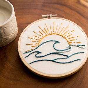 Peut inclure: Art de broderie en forme de cercle repr&eacute;sentant un soleil jaune avec des rayons et des vagues bleues sur un fond blanc.