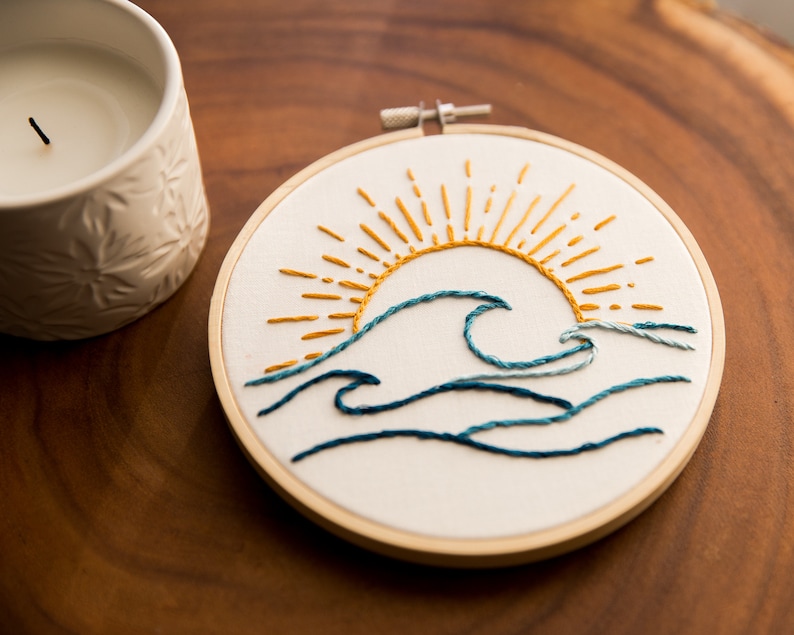 Peut inclure: Art de broderie en forme de cercle repr&eacute;sentant un soleil jaune avec des rayons et des vagues bleues sur un fond blanc.