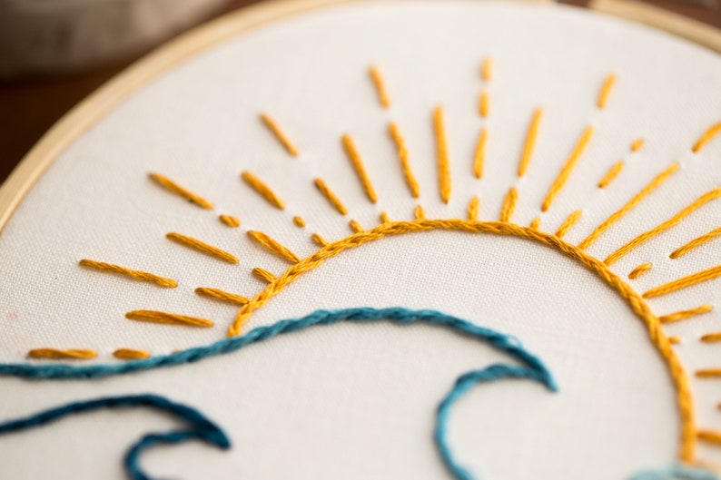 Peut inclure: Gros plan sur un motif de broderie fait main repr&eacute;sentant un soleil jaune avec des rayons et une vague bleue.