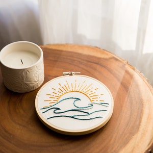 Peut inclure: Un cercle de broderie blanc avec un motif brod&eacute; &agrave; la main d'un soleil jaune se levant au-dessus de vagues bleues. Le cercle est sur une surface en bois &agrave; c&ocirc;t&eacute; d'une bougie blanche.
