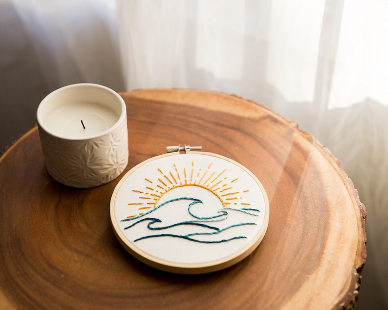 Peut inclure: Un cercle de broderie blanc avec un motif brod&eacute; &agrave; la main d'un soleil jaune se levant au-dessus de vagues bleues. Le cercle est sur une surface en bois &agrave; c&ocirc;t&eacute; d'une bougie blanche.