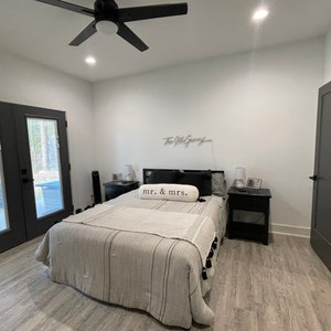 May include: A bedroom with a bed, a ceiling fan, and a door. The bed has a white and grey striped duvet and a white pillow with the text "mr. & mrs." on it. There are two bedside tables on either side of the bed, each with a lamp and a small picture frame.