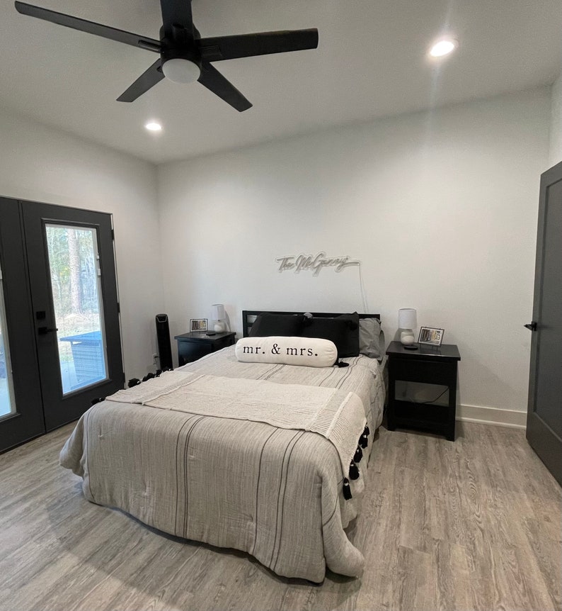 May include: A bedroom with a bed, a ceiling fan, and a door. The bed has a white and grey striped duvet and a white pillow with the text "mr. & mrs." on it. There are two bedside tables on either side of the bed, each with a lamp and a small picture frame.