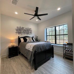 May include: A bedroom with a black bed frame, grey bedding, and a black and white geometric wall art piece. The room has a ceiling fan, a window with a view of trees, and a wooden floor.