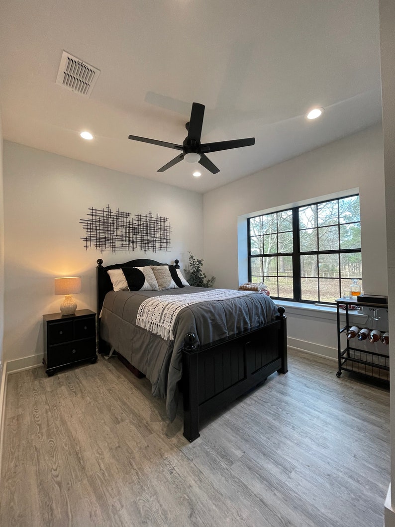 May include: A bedroom with a black bed frame, grey bedding, and a black and white geometric wall art piece. The room has a ceiling fan, a window with a view of trees, and a wooden floor.