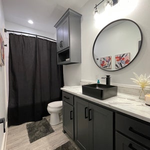 May include: A modern bathroom with a black shower curtain, a white toilet, a black sink with a white countertop, and grey cabinets. The bathroom has a black framed round mirror and a light fixture with three glass bulbs.
