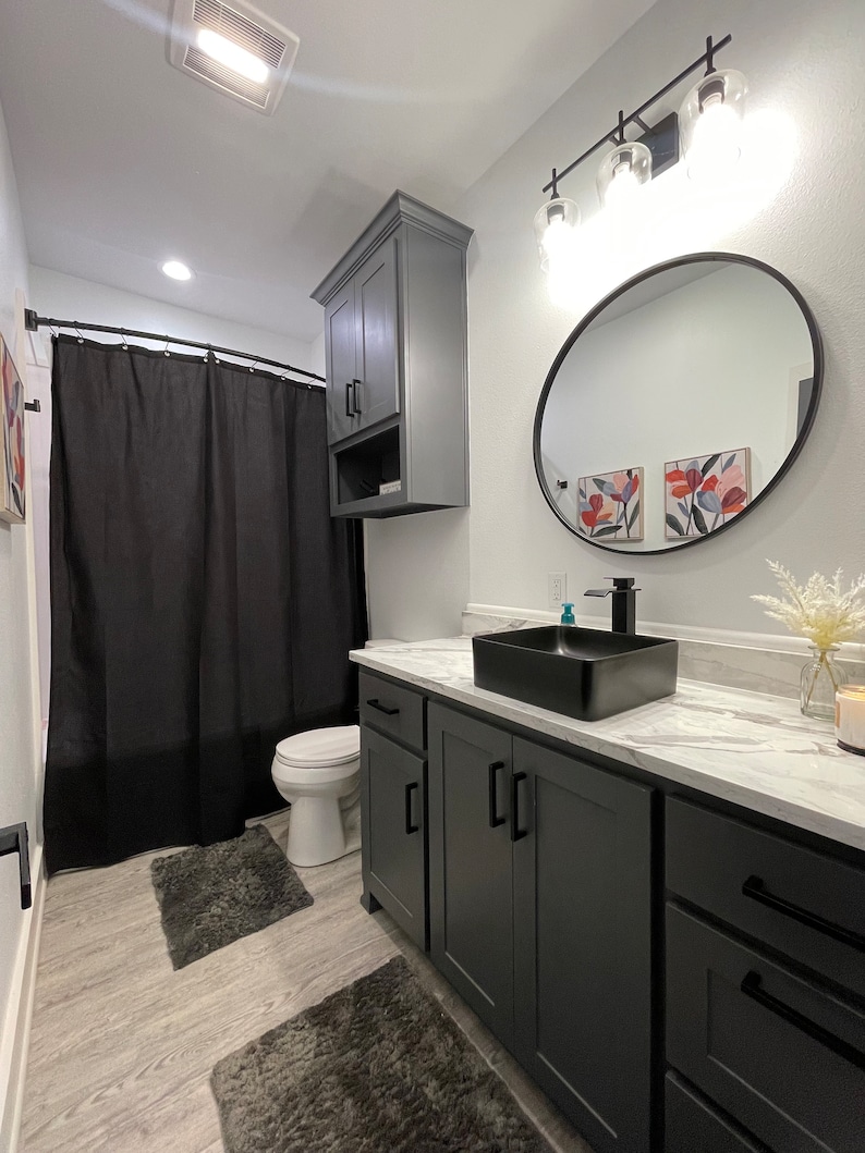 May include: A modern bathroom with a black shower curtain, a white toilet, a black sink with a white countertop, and grey cabinets. The bathroom has a black framed round mirror and a light fixture with three glass bulbs.
