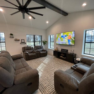 May include: A living room with a large flat screen TV mounted on the wall, a brown leather sofa, two brown leather recliners, and a black TV stand. The room has large windows with black frames and a ceiling fan.