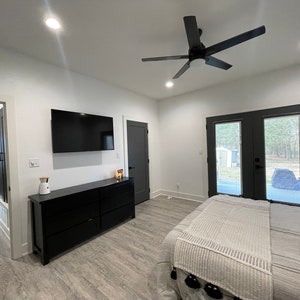 May include: A bedroom with a white bed, a black dresser, a flat screen TV, and a ceiling fan. The room has a white wall and a grey floor. There are two black doors leading to the outside.