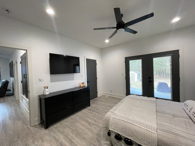 May include: A bedroom with a white bed, a black dresser, a flat screen TV, and a ceiling fan. The room has a white wall and a grey floor. There are two black doors leading to the outside.