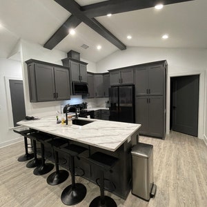 May include: A modern kitchen with dark grey cabinets, a white marble countertop island, and black bar stools. The island has a stainless steel bin and a sink with a tap.
