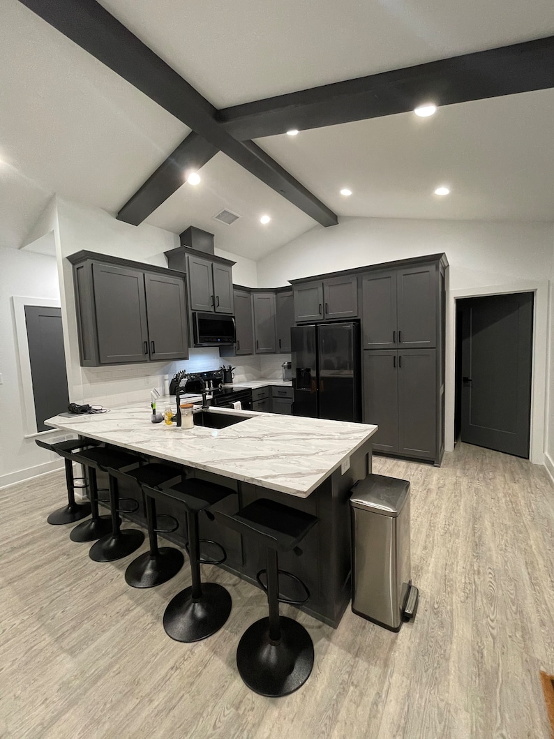 May include: A modern kitchen with dark grey cabinets, a white marble countertop island, and black bar stools. The island has a stainless steel bin and a sink with a tap.