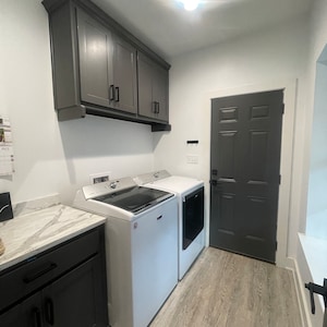 May include: A white laundry room with a grey door, grey cabinets, and two white washing machines. The floor is a light wood laminate.