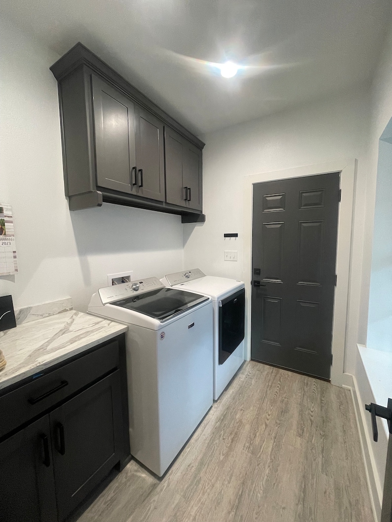 May include: A white laundry room with a grey door, grey cabinets, and two white washing machines. The floor is a light wood laminate.