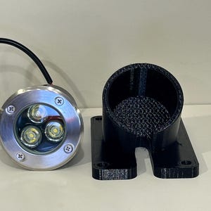 Könnte beinhalten: Ein LED-Strahler aus Edelstahl mit drei hellen Lichtern und einem schwarzen Kabel. Daneben befindet sich ein schwarzer, 3D-gedruckter zylindrischer Halter mit rechteckiger Basis. Die Artikel sind wahrscheinlich für die Außenbeleuchtung.