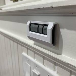 May include: A white wall-mounted switch with three buttons. The buttons are gray and have a raised design. The switch is mounted on a white wall.
