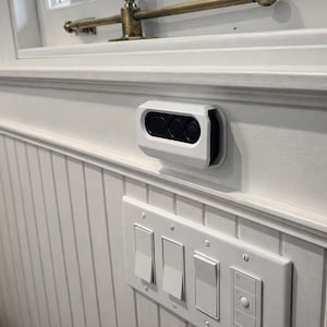 May include: White wall-mounted smart home device with three black circular lenses. Below, a white light switch panel with four switches is mounted on a white paneled wall. The scene is illuminated by natural light.
