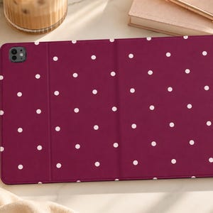 May include: A rectangular tablet case in a deep burgundy color featuring a repeating pattern of small white polka dots. The case has a smooth, matte finish and includes a dedicated cutout for a rear-facing camera lens on the top left corner.