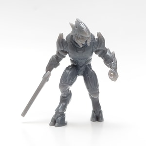 Peut inclure: Figurine en plastique gris d'un extraterrestre humanoïde avec une grosse tête et une arme à la main.