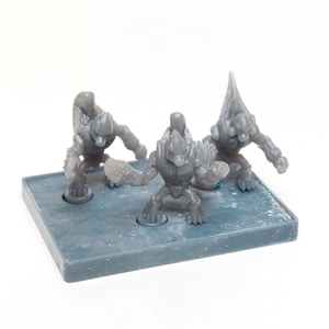 Peut inclure: Trois figurines grises représentant des créatures extraterrestres sur une base bleu-gris. Les figurines ont une armure détaillée et sont dans des poses dynamiques. La base a des indentations circulaires.