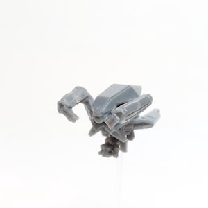 May include: A small, grey, plastic toy robot with articulated arms and legs. The robot has a geometric design with a central body and multiple appendages. The toy is set against a plain white background.