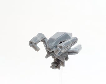 Pode incluir: Um pequeno robô de brinquedo de plástico cinza com braços e pernas articulados. O robô tem um design geométrico com um corpo central e múltiplos apêndices. O brinquedo está sobre um fundo branco liso.
