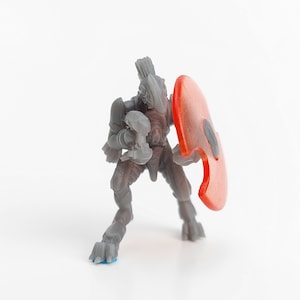 Può includere: Action figure grigia di un guerriero futuristico, con un'arma e uno scudo rosso. La figura è in posa dinamica, con armatura dettagliata e un elmo. Lo scudo ha un design ritagliato unico.