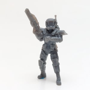 Könnte beinhalten: Graue Miniaturfigur eines futuristischen Soldaten, der eine große Waffe hält. Die Figur ist detailliert mit Rüstung, Helm und Taschen. Das Gesamtdesign deutet auf ein Science-Fiction- oder Militärthema hin.