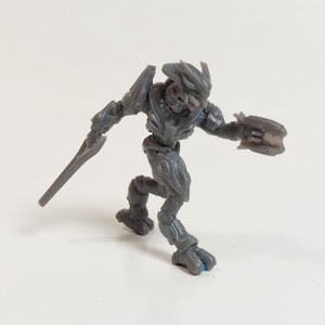 May include: A gray action figure of a humanoid alien in a dynamic pose, holding a blade in one hand and a weapon in the other. The figure has detailed armor and is set against a white background.