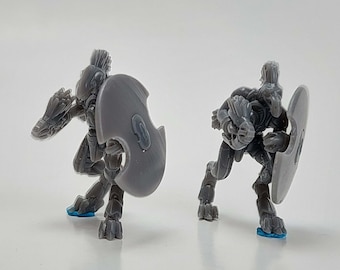 May include: Two gray miniature robot figures in a defensive stance, each holding a shield. The robots have detailed armor and are posed on a white surface. The bases are blue.