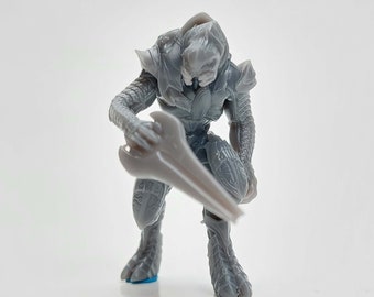 May include: A gray action figure of an alien warrior, holding a large, curved blade. The figure is detailed with armor and clawed feet, posed on a white surface. The figure is approximately 4 inches tall.