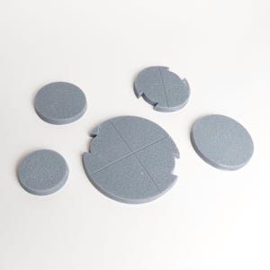 May include: Five gray plastic round bases for miniature figures. The bases are different sizes and some have a cross pattern.