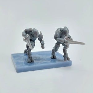 May include: Two gray plastic action figures of alien warriors holding energy weapons. The figures are standing on a gray plastic base.