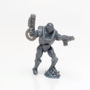 Op de afbeelding: Een grijze miniatuurfiguur van een futuristische soldaat, gewapend met twee geweren en gekleed in een gedetailleerd pantser. De figuur staat in een actiehouding, met één arm omhoog die een groot wapen vasthoudt. De basis van de figuur is getextureerd met gekartelde spikes.