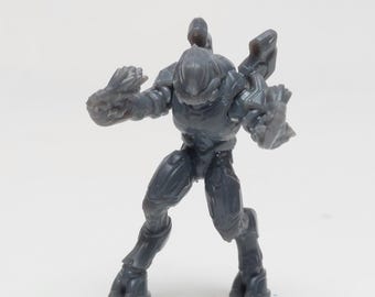 May include: A gray plastic action figure of a sci-fi alien warrior. The figure is posed in a dynamic fighting stance with its arms raised and its legs spread apart. The figure has a detailed sculpt with intricate armor and weapons.