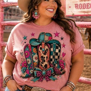 May include: A dusty rose t-shirt featuring a graphic print of three cowboy boots, a cowboy hat, and a bow. The boots display floral, leopard, and checkered patterns in shades of turquoise, pink, and orange. Small stars and hearts surround the design.