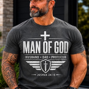 May include: A charcoal grey short-sleeved t-shirt featuring white distressed text that reads MAN OF GOD, HUSBAND, DAD, PROTECTOR, and JOSHUA 24:15. The graphic includes a cross, a shield with a sword, and wing-like accents centred on the chest.