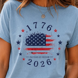 Pode incluir: Camiseta azul clara com um design desgastado da bandeira americana, o ano de 1776 e o ano de 2026. A camiseta também inclui o texto "250 YEARS OF FREEDOM" e é rodeada por estrelas.