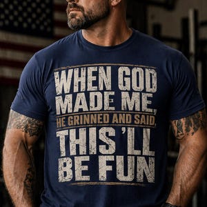 May include: Navy blue t-shirt with the text "WHEN GOD MADE ME, HE GRINNED AND SAID, THIS'LL BE FUN" in a distressed white font. The shirt is worn by a person wearing a black cap and watch.
