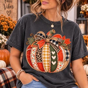 Może przedstawiać: Ciemnoszary t-shirt z krótkim rękawem z dużą grafiką przedstawiającą dynię złożoną z różnych materiałowych łat. Wzór dyni zawiera motywy takie jak panterka, żółta kratka, czerwone kwiaty oraz czarna krata.