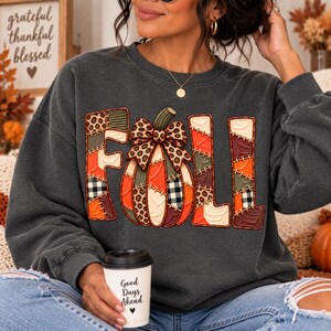 May include: A charcoal gray crewneck sweatshirt featuring the word FALL in large, patchwork-style letters. The letter A is replaced by a pumpkin with a leopard print bow. The design incorporates orange, brown, green, and black and white plaid patterns.