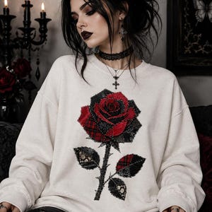 May include: An off-white long-sleeve sweatshirt featuring a large, central rose graphic. The rose is constructed from layered fabric patches in red and black, including plaid and lace textures, with visible stitching along the edges of the petals and leaves.