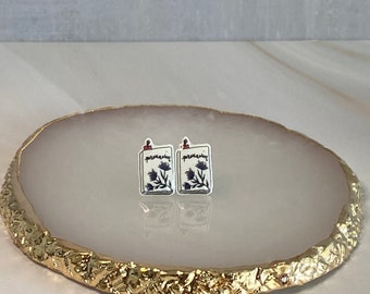 May include: A pair of small, white, book-shaped earrings with a floral design and the word "remarkable" printed on them. The earrings are on a light pink and gold agate coaster.