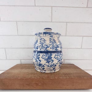 May include: A ceramic jar with a lid, featuring a cream-colored base and a blue splatter pattern. The jar is cylindrical with a slightly wider base and a matching lid. It sits on a wooden surface against a white brick wall.