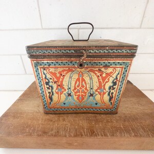 K&ouml;nnte beinhalten: Eine antike, quadratische Blechdose mit einem Scharnierdeckel und einem Metallgriff. Die Dose ist mit einem farbenfrohen, kunstvollen Design verziert, das orange, blaue und goldene Akzente aufweist. Das Design erinnert an traditionelle islamische Kunst.