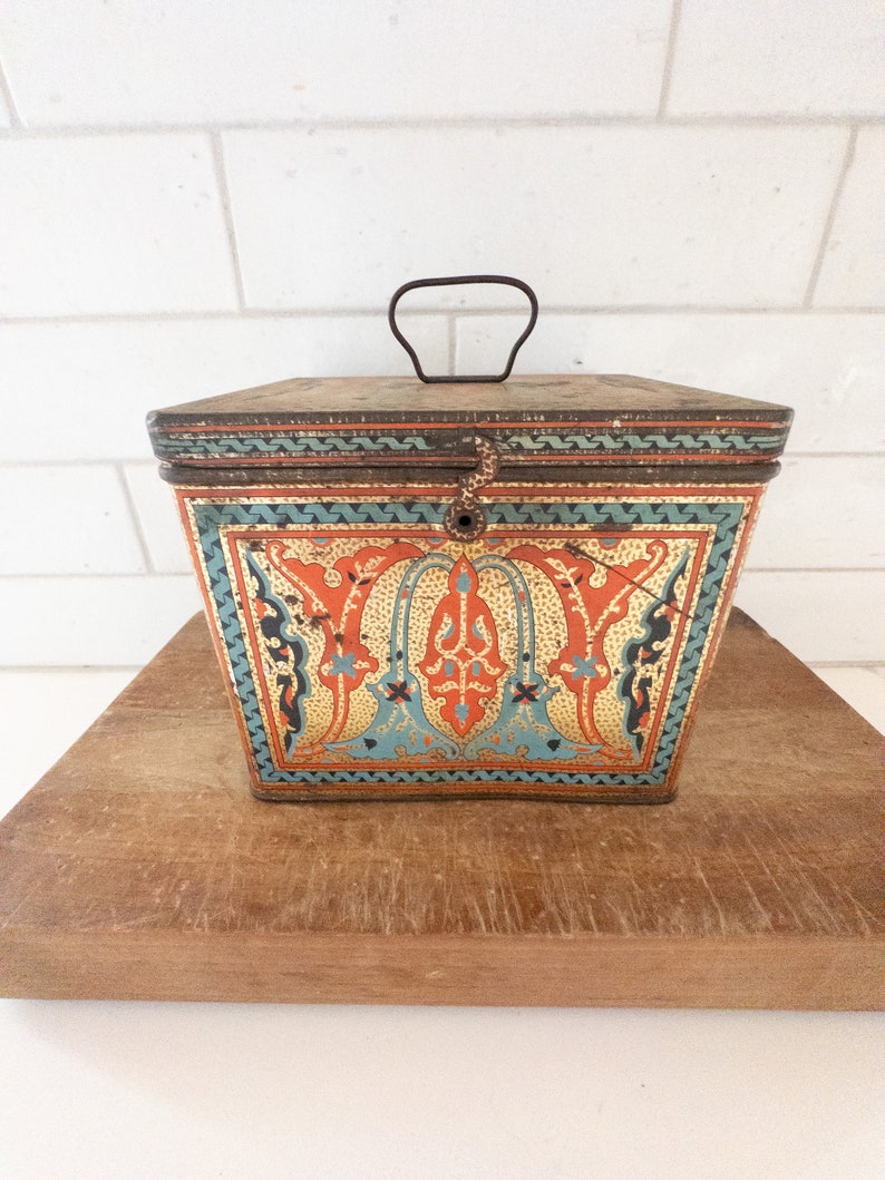 K&ouml;nnte beinhalten: Eine antike, quadratische Blechdose mit einem Scharnierdeckel und einem Metallgriff. Die Dose ist mit einem farbenfrohen, kunstvollen Design verziert, das orange, blaue und goldene Akzente aufweist. Das Design erinnert an traditionelle islamische Kunst.