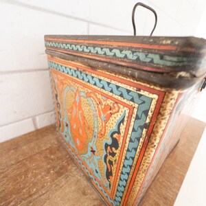 K&ouml;nnte beinhalten: Eine vintage Metalldose mit einem dekorativen Design mit komplizierten Mustern in Orange-, Blau- und Goldt&ouml;nen. Die Dose hat einen Klappdeckel und einen Drahtgriff.